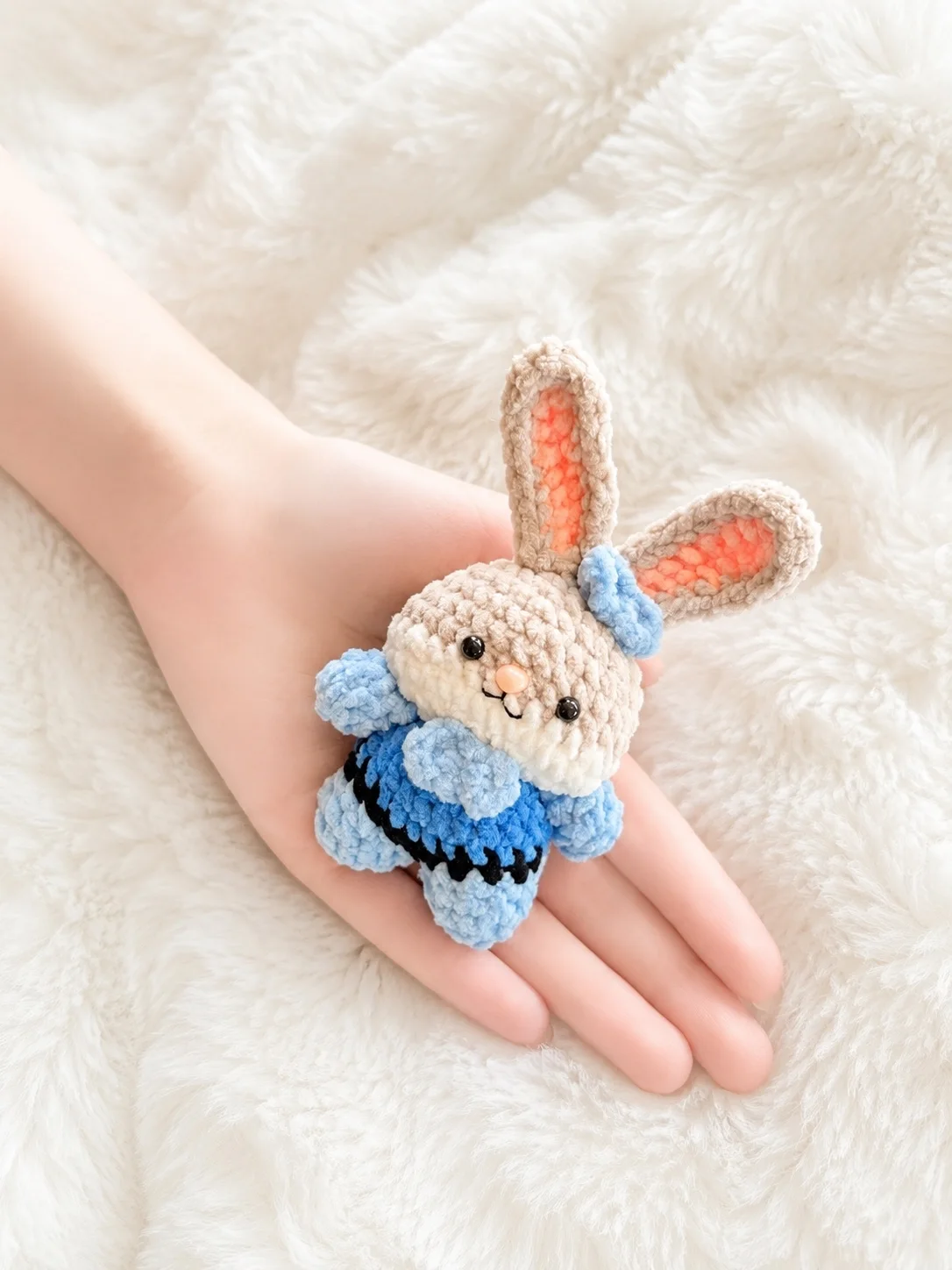 Beige crochet bunny with upright pink-lined ears and a blue outfit, resting in a hand