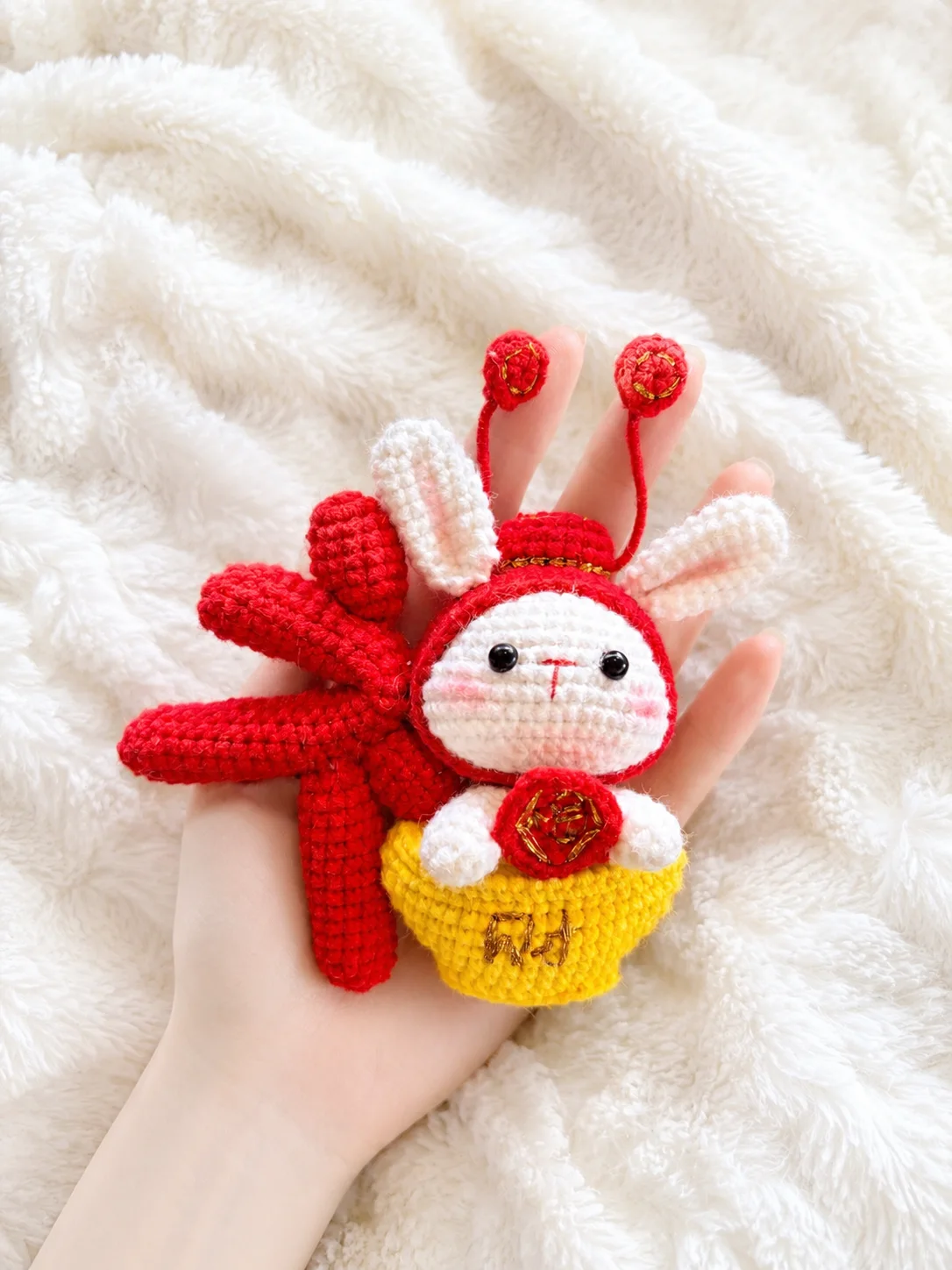 White crochet bunny in a red festive costume with gold details, a yellow ingot-shaped base and red hanging decorations, held in a hand