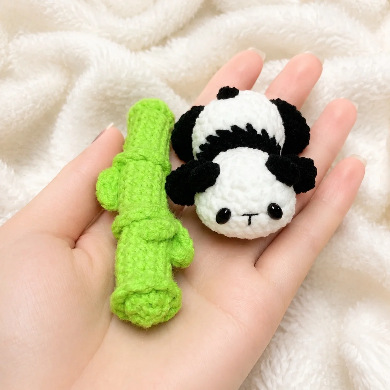 Small black and white crochet panda lying beside a bright green crochet bamboo stalk in a palm