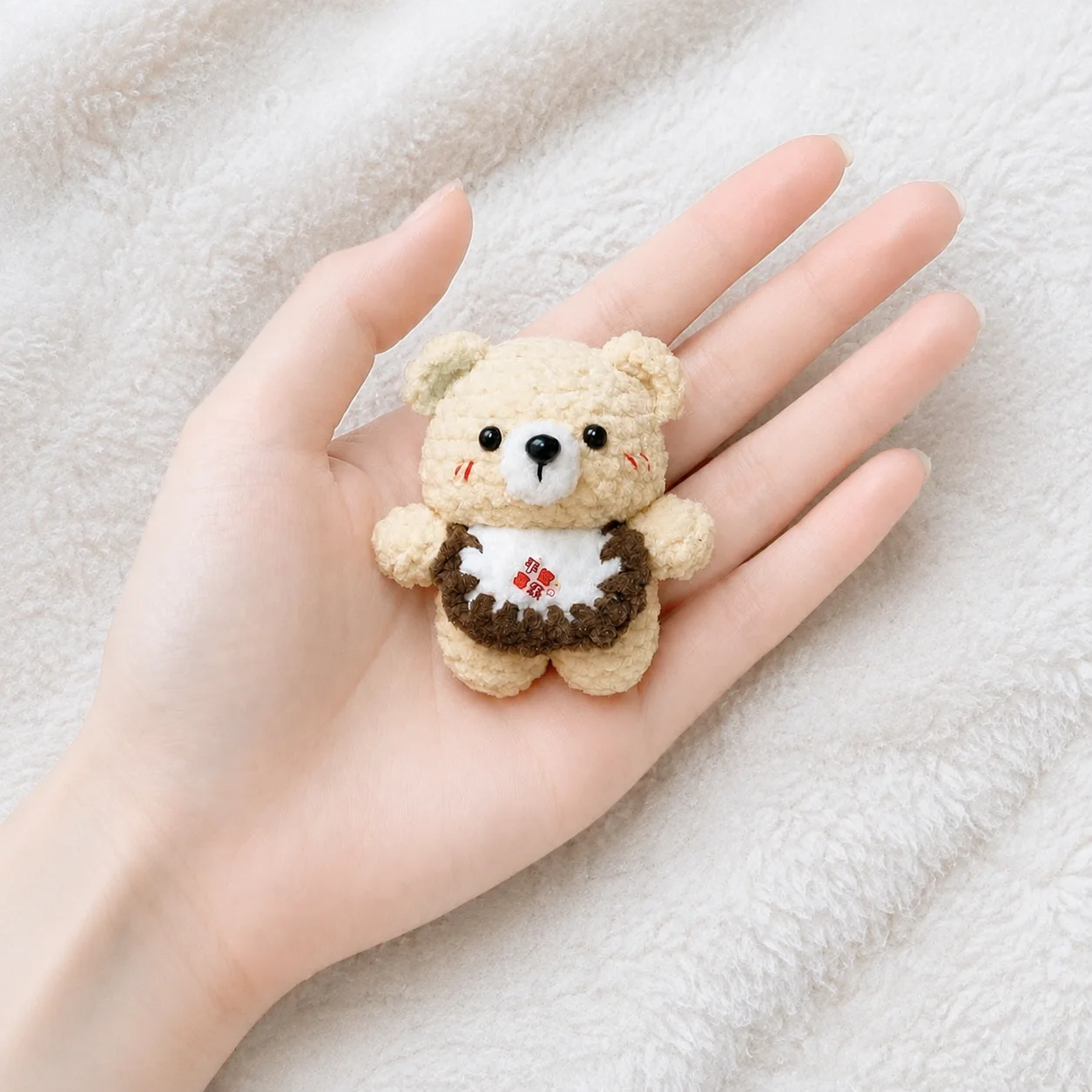 Tiny golden-beige crochet bear with a white muzzle and a brown-edged bib, shown in a hand