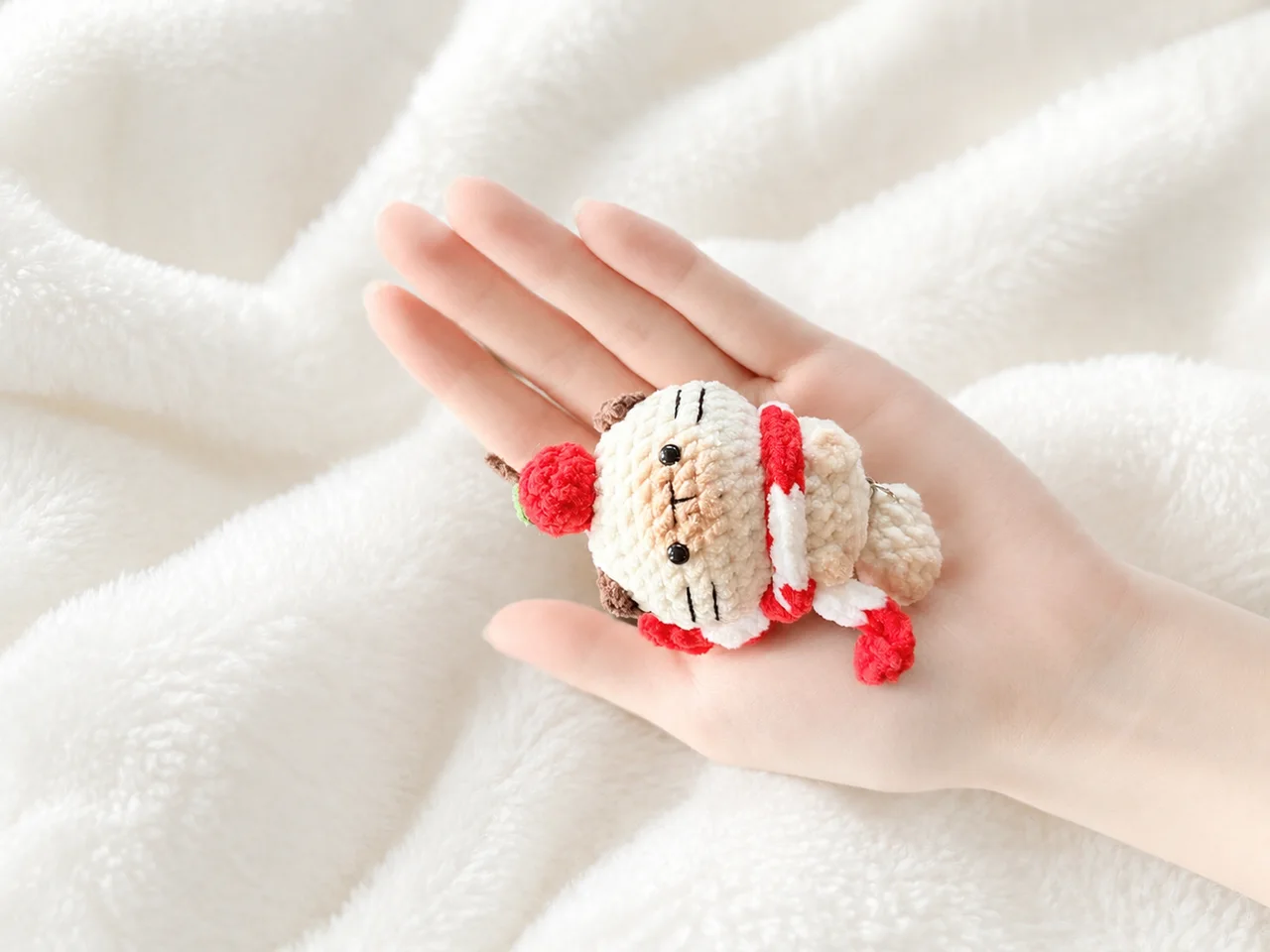 Tiny cream crochet cat with brown ears, stitched whiskers, a red fruit on its head and a red-and-white scarf, resting in a hand