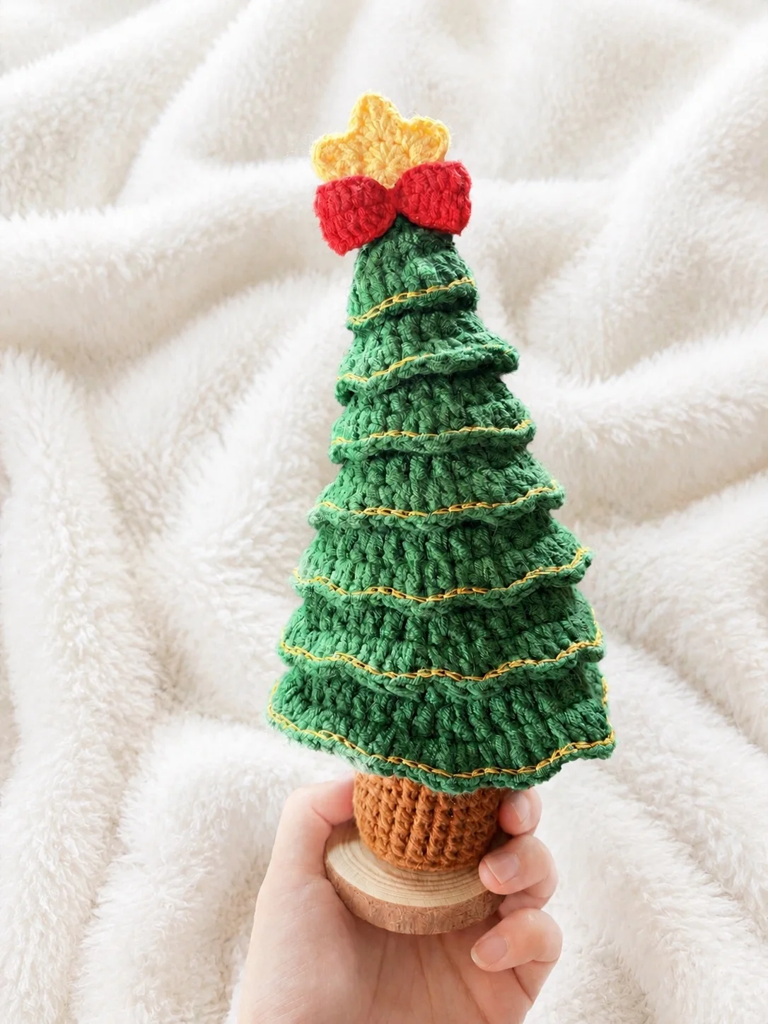 Green crochet Christmas tree with gold edging, a red bow and a yellow star on a wooden base, held in a hand