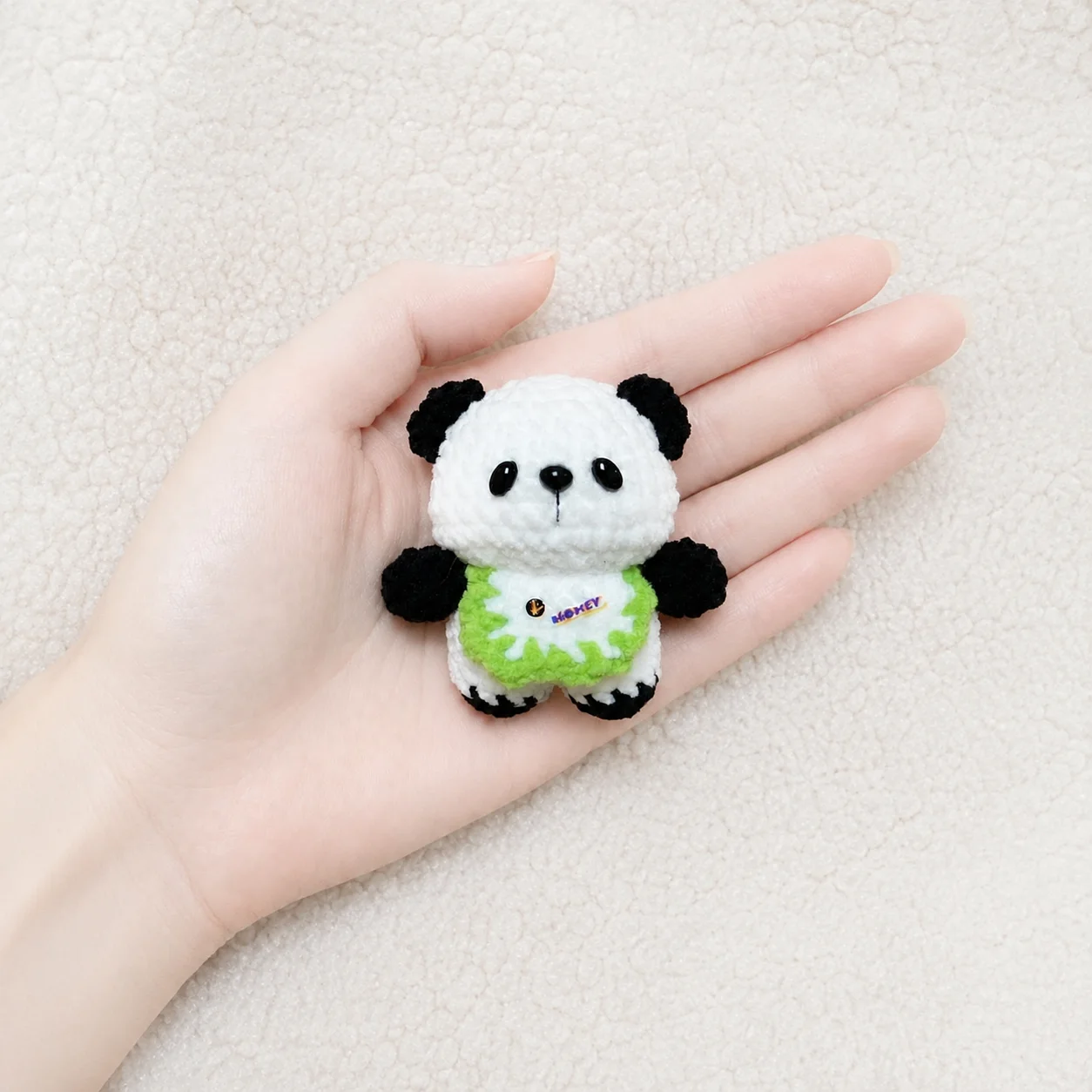 Tiny upright black-and-white crochet panda wearing a bright-green-edged white bib, resting in a palm