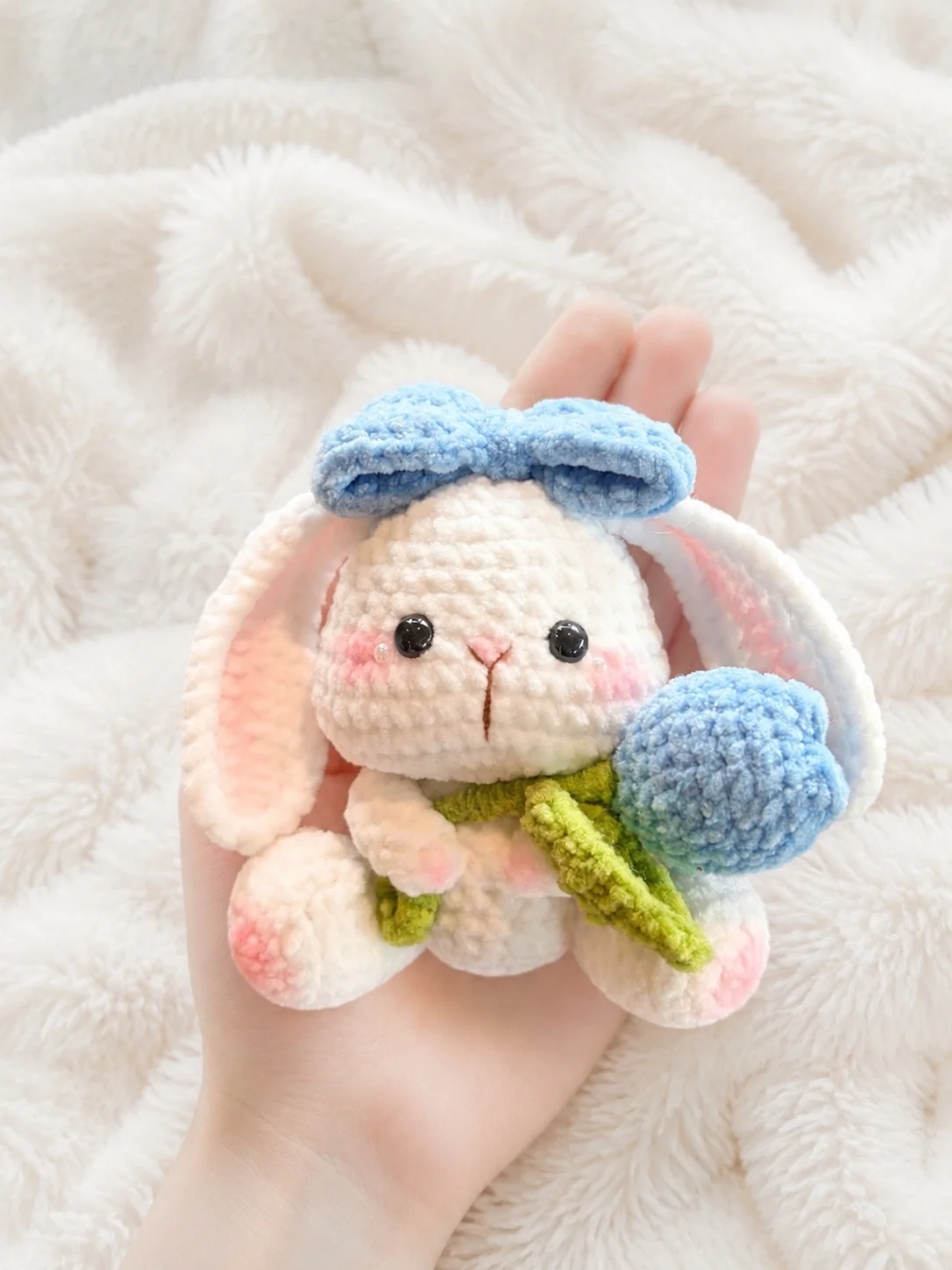 A white crochet bunny with a blue bow holding a blue tulip, resting in a hand