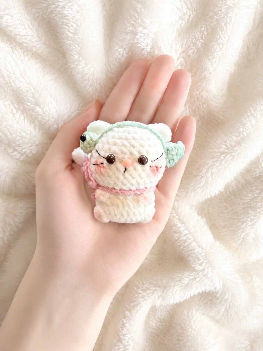 Small white crochet kitten wearing mint-green headphones and a pink shoulder bag, held in a hand