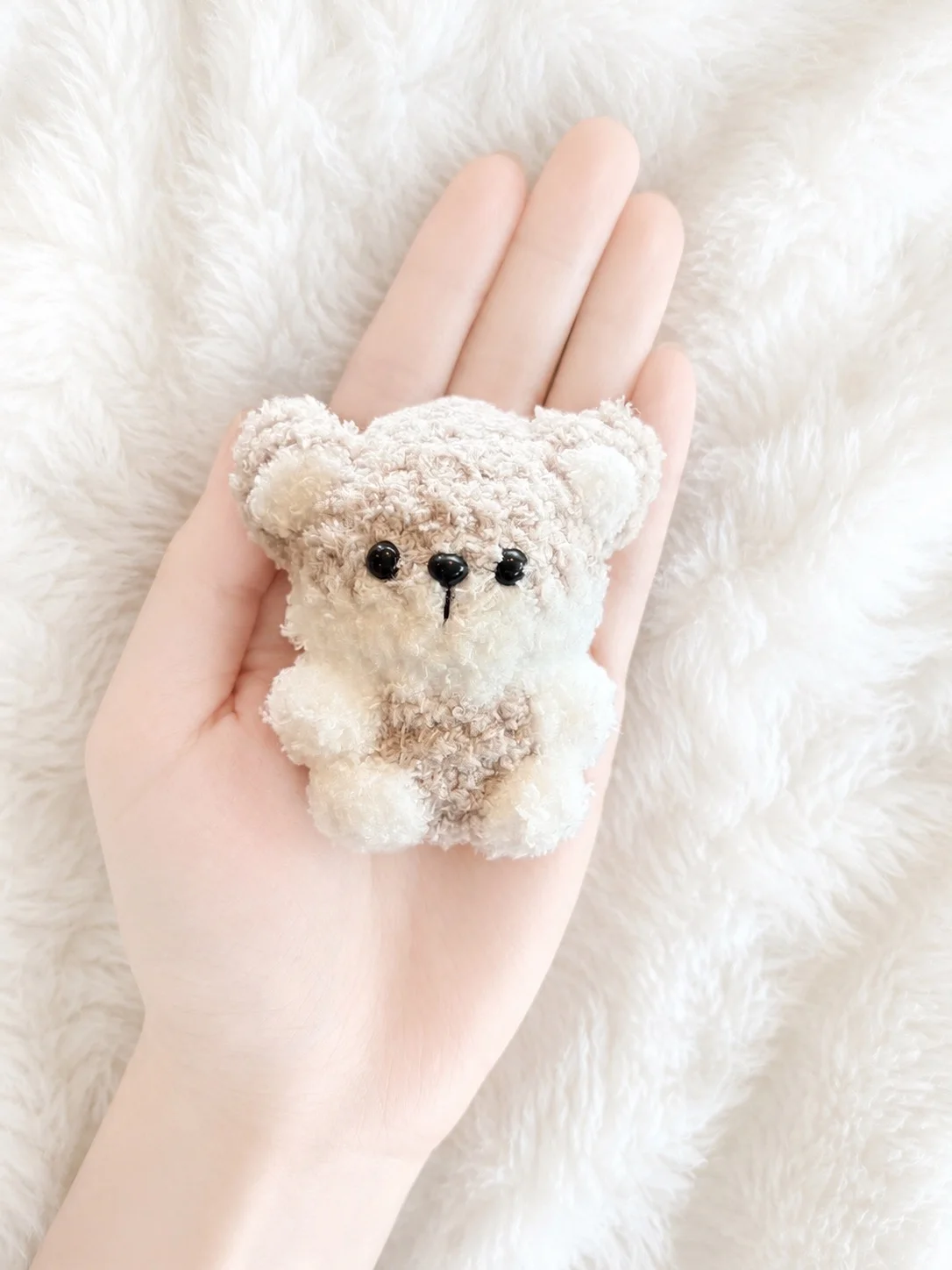 Tiny fluffy cream-and-taupe crochet bear with round ears, a dark nose and a taupe tummy, resting in a hand