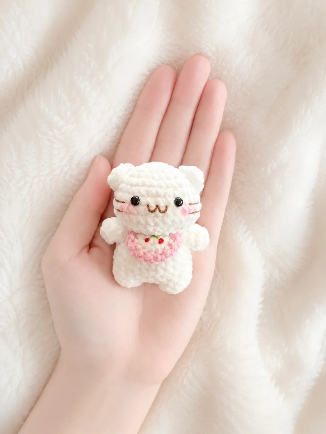 Tiny white crochet kitten with stitched whiskers and a pink-edged bib decorated with cherries, held in a hand