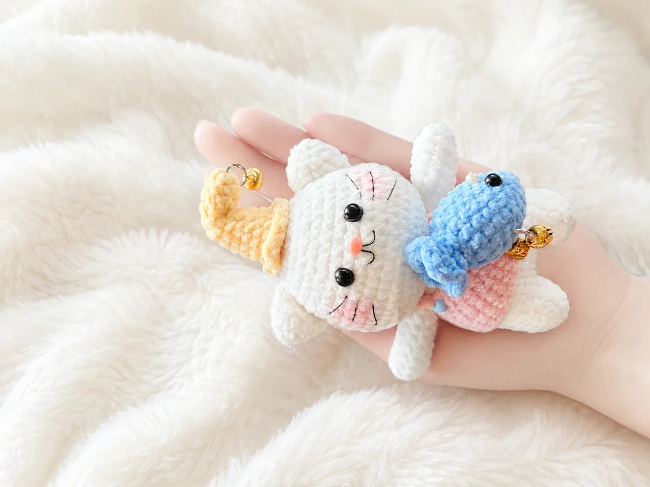 White crochet cat in a curled yellow hat and pink outfit, holding a blue fish with little gold bells, resting in a hand