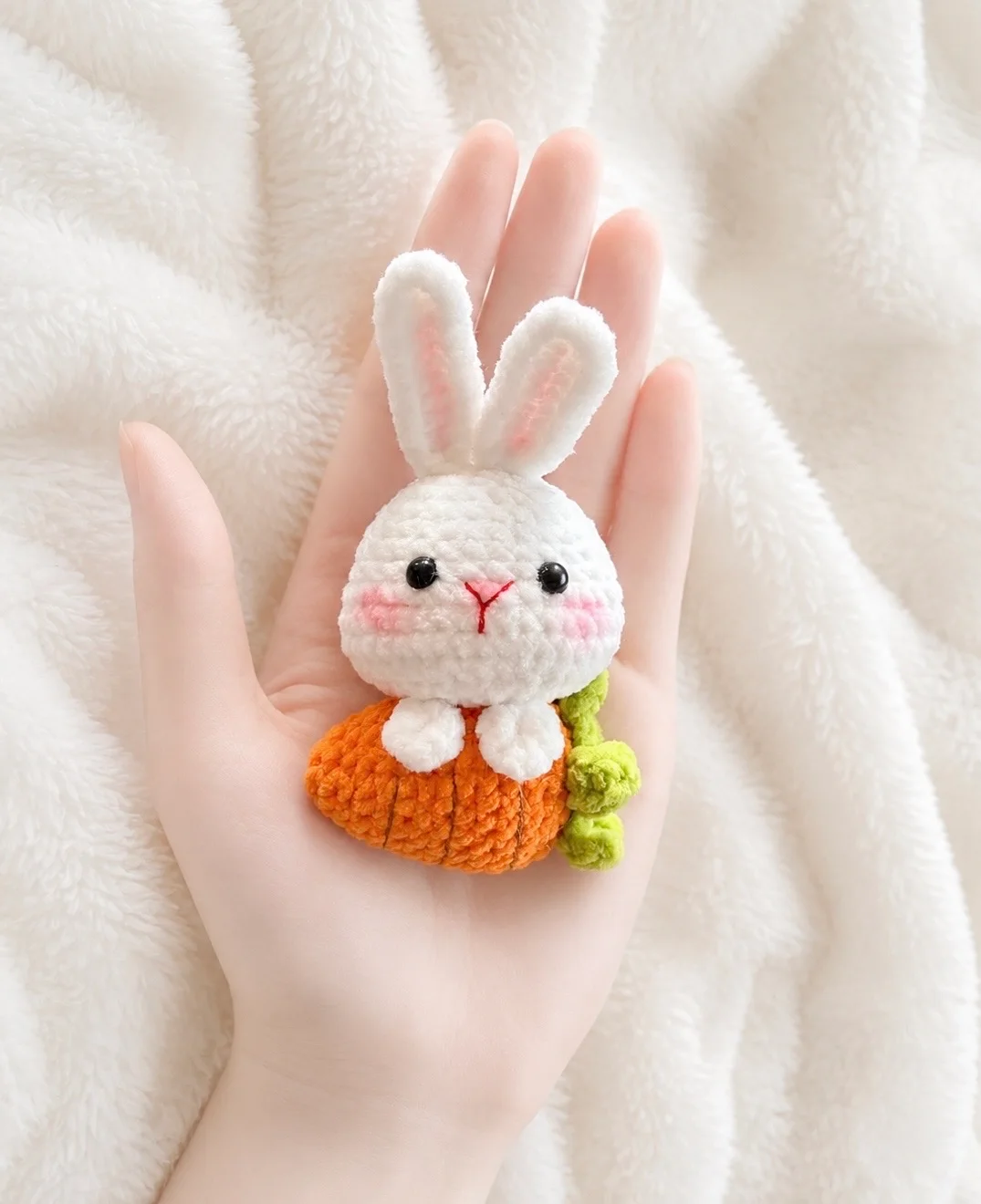 White upright-eared crochet bunny nestled in an orange carrot with green leaves, shown in a hand
