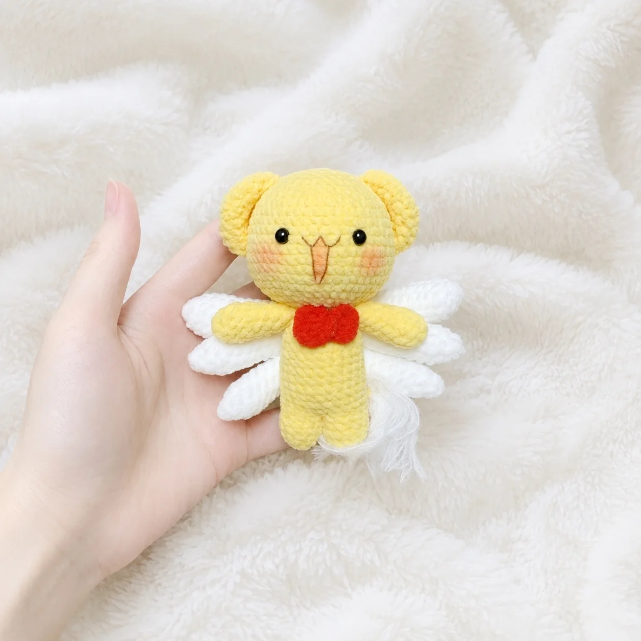 Yellow crochet character with round ears, white wings and a red bow tie, held in a hand