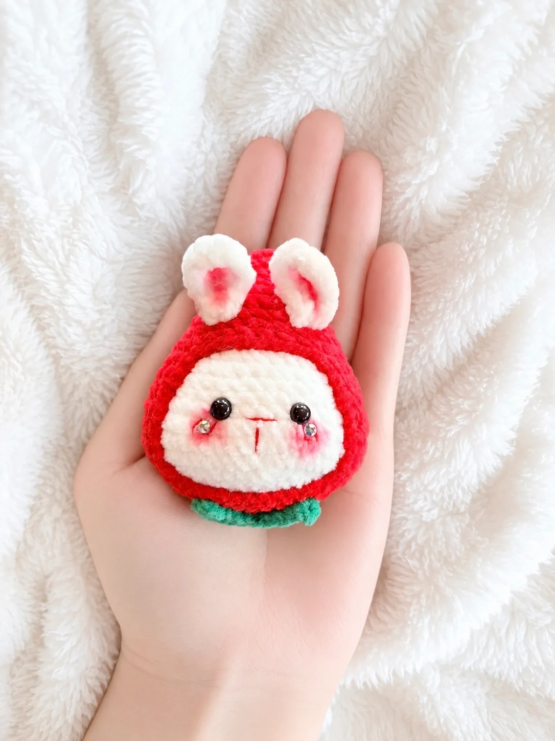 White crochet bunny peeking out of a red strawberry costume with green leaves, shown from the front in a hand