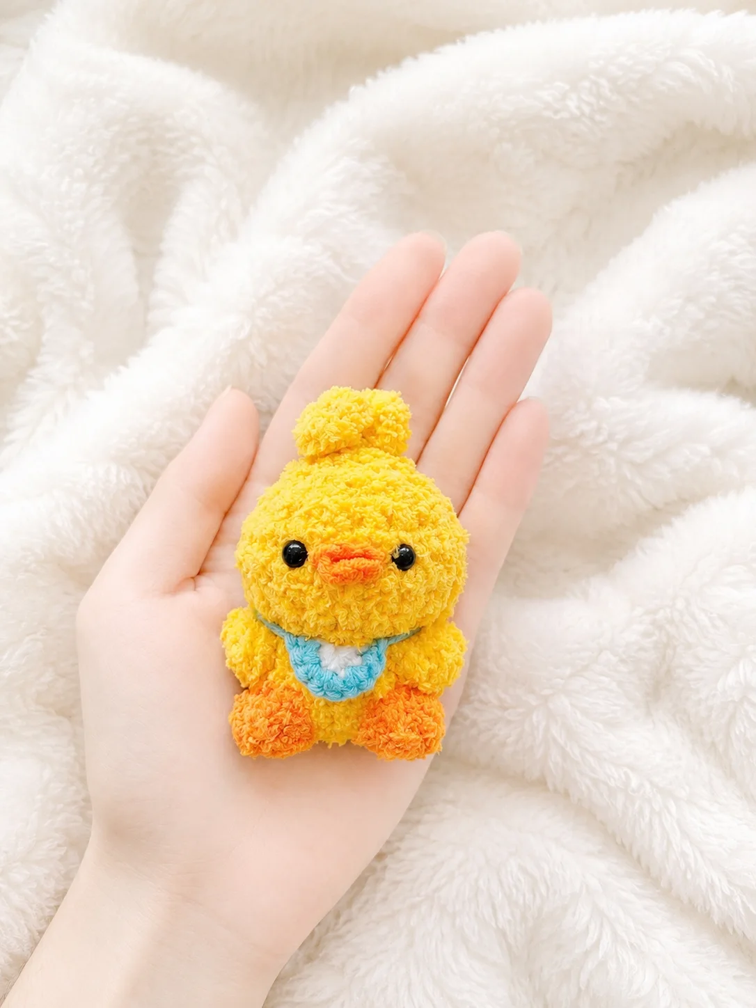 Fluffy yellow crochet duckling with an orange beak and feet and a blue-and-white bib, resting in a hand