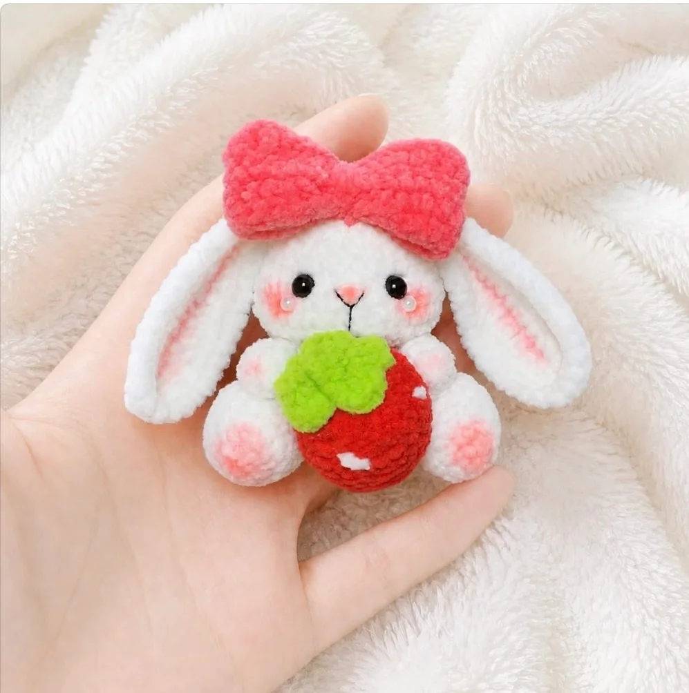 White floppy-eared crochet bunny with a coral-pink bow holding a red strawberry with green leaves, resting in a hand