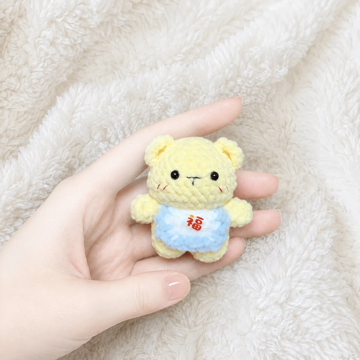 Tiny pale-yellow crochet bear wearing a light-blue bib with a red Chinese character, resting in a hand