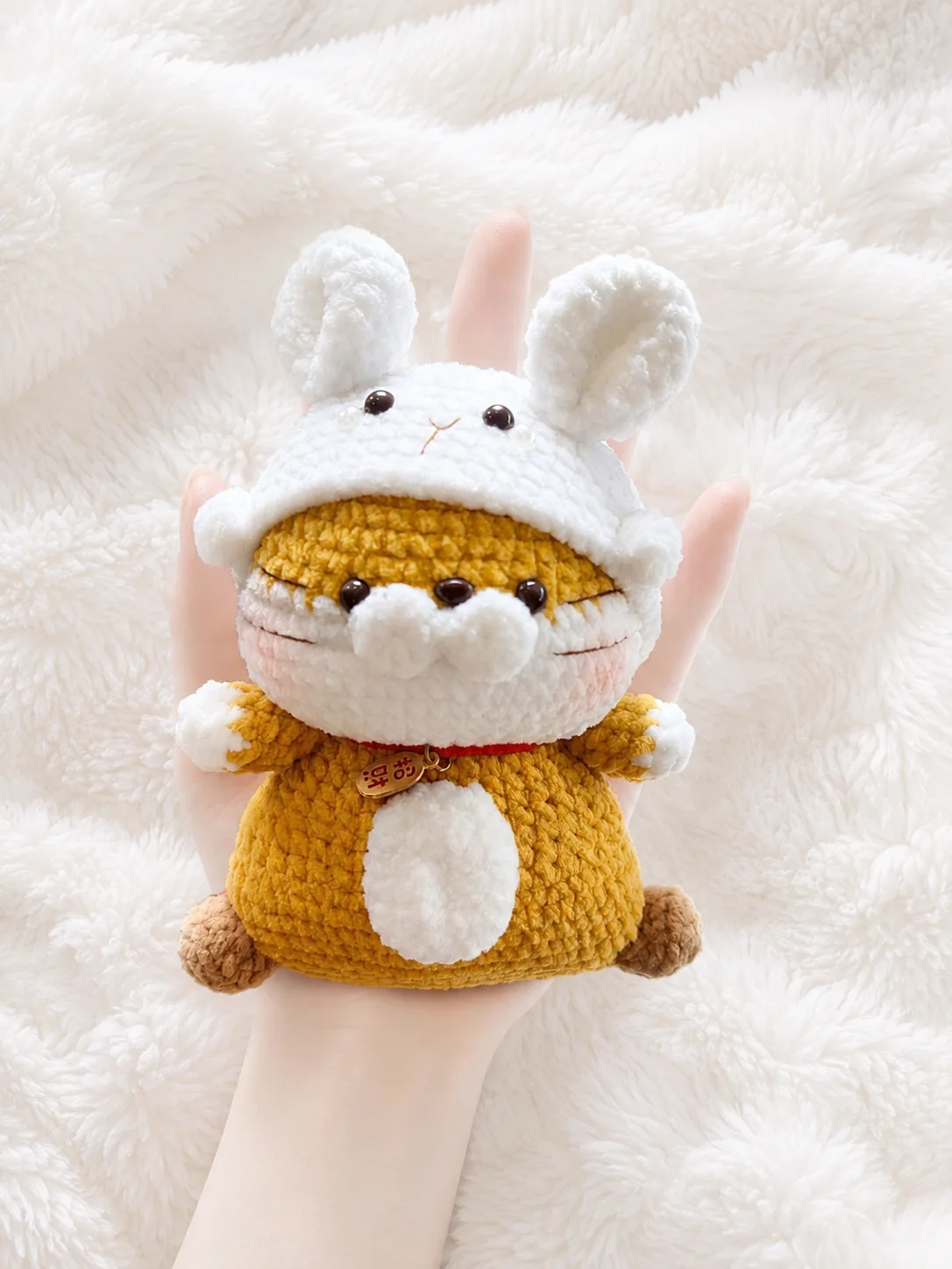 Golden crochet tiger with a cream muzzle and tummy, wearing a white bunny hat with tall ears and a red collar, held in a hand